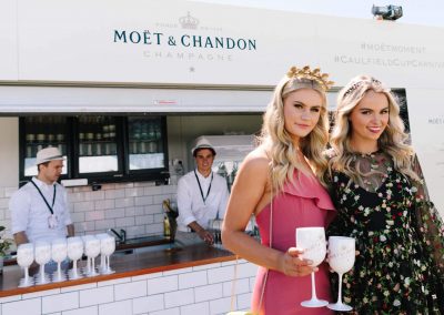 2 female models holding white acrylic champagne goblet glasses bar and 2 male bartenders background